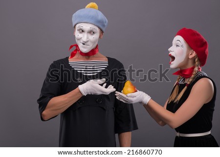 Waist-up portrait of mime couple with white faces, female mime giving a yellow pear to male mime looking on her isolated on grey background with copy place