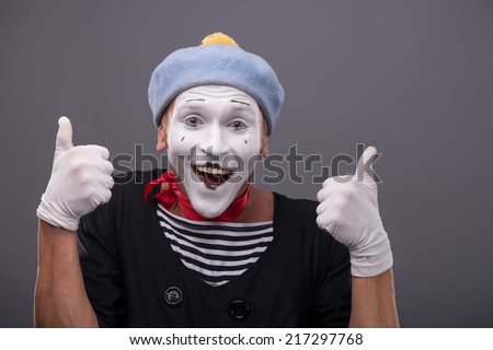 Waist-up portrait of funny male mime with grey hat and white face showing sign OK with both hands and looking at the camera with happy smile isolated on grey background with copy place