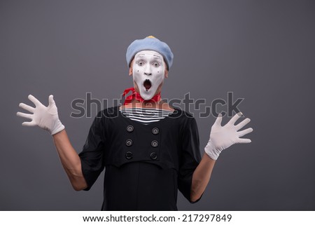 Waist-up portrait of funny male mime with grey hat and white face looking at the camera with great surprise, opening his mouth and breeding his hands isolated on grey background with copy place