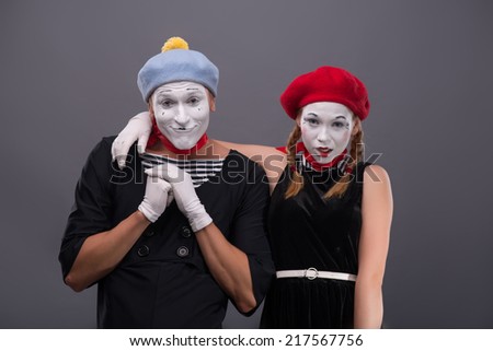 Waist-up portrait of funny mime couple looking at the camera, male mime holding his hands on the chest, female mime hugging him isolated on grey background with copy place