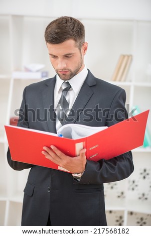 Waist-up portrait of handsome confident businessman standing in office with a red folder in his hands and reading with serious face
