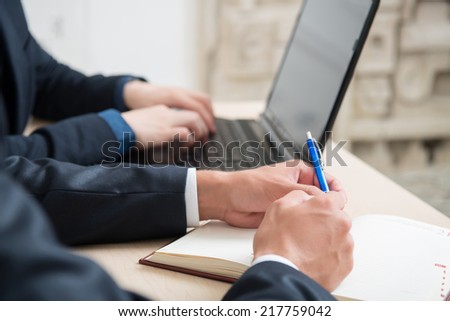 Close-up photo of hands of businessmen in suits sitting at the table with laptop in office interior, one man typing and another man writing with blue pen in his notebook