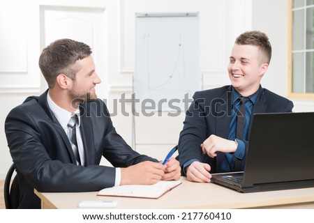 Waist-up portrait of two handsome businessmen in suits sitting at the table with laptop in office interior, laughing and looking on each other while discussing a new project
