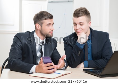Waist-up portrait of two handsome businessmen in suits sitting at the table in office interior and discussing something that one have showed another on his mobile phone