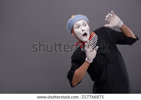 Waist-up portrait of young male mime confused breeding his hands showing that he is scared and frighten looking aside with wide opened mouth and eyes isolated on grey background with copy place