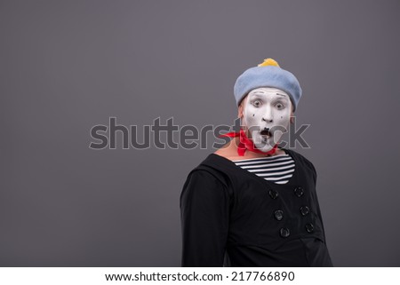 Waist-up Portrait of young male mime with white face, grey hat opening his mouth and looking aside with astonishment isolated on grey background with copy place