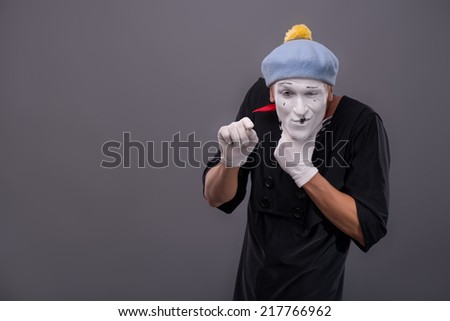 Waist-up Portrait of young male mime with white face, grey hat insidiously scheming something and looking at the camera isolated on grey background with copy place