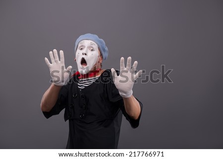 Waist-up Portrait of young male mime confused breeding his hands showing that he is scared and frighten looking at you with wide opened mouth and eyes isolated on grey background with copy place