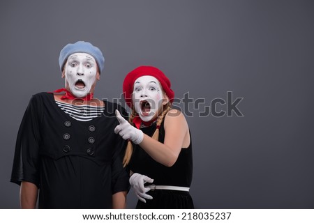 Waist-up portrait of scared mime couple looking at the camera, female mime loudly crying and showing at you standing near male mime with startled face isolated on grey background with copy place