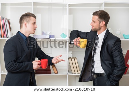 Waist-up portrait of two handsome businessmen in suits looking on each other while having a coffee-break in office standing near a bookshelf and discussing something