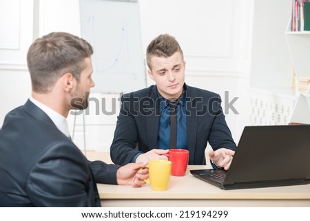 Waist-up portrait of two handsome businessmen in suits having a coffee-break in office sitting at the table, with selective focus on one showing his colleague something in his laptop