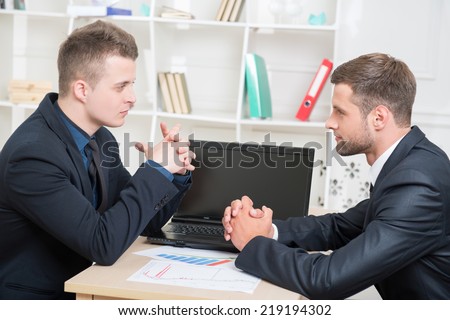 Waist-up portrait of two angry businessmen in suits sitting at the table in office looking on each other with spite thinking about some problems