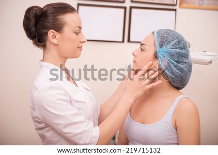Waist-up portrait of a cosmetologist inspecting skin of her patient a young woman with fresh and clean skin seriously looking at her after a professional cosmetology procedures in a beauty salon