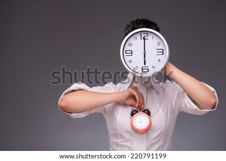 Close-up portrait of girl holding in one hand a big clock and in other hands small alarm clock, hiding her face with a big clock, isolated on grey background with copy place