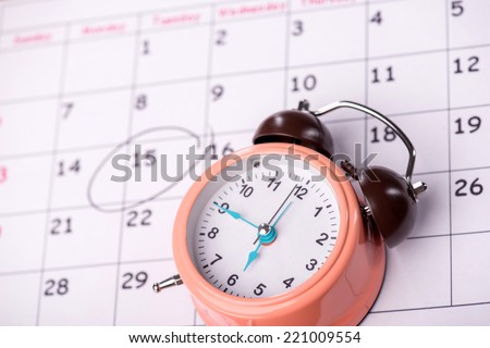 Close-up top-view photo of calendar with a datum circled with a black marker, with selective focus on an alarm clock, concept of time management at work