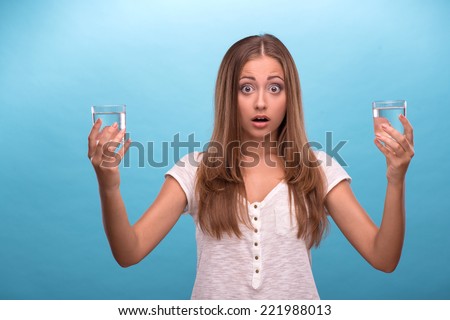 Waist-up portrait of young beautiful girl with confused face holding two glasses with clean clear water looking at the camera comparing water in glasses isolated on blue background with copy place