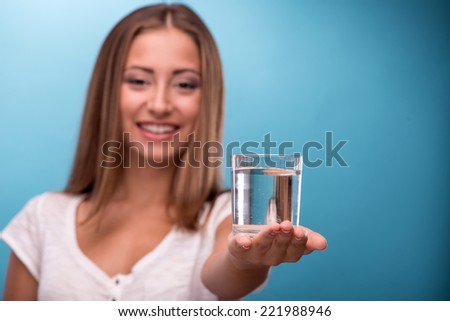 Waist-up portrait of young beautiful girl with confused face holding two glasses with clean clear water looking at the camera comparing water in glasses isolated on blue background with copy place