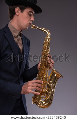 Waist-up portrait of a young handsome Caucasian jazz man in a suit with a black hat looking at the camera with astonishment holding a trumpet in his hand isolated on grey background with copy place