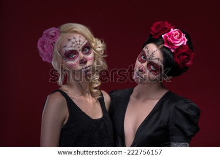 Waist-up portrait of two young girls standing near each other in black dresses with Calaveras makeup and roses in their hair looking at the camera isolated on red background with copy place