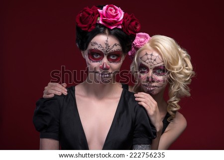 Waist-up portrait of two young girls in black dresses with Calaveras makeup and roses in their hair looking at the camera, one stands behind another isolated on red background with copy place