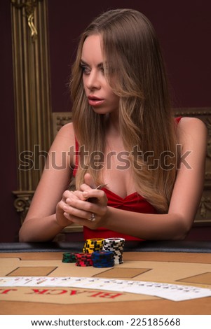 Beautiful woman playing in casino holding in hands chips and cards sitting at table looking aside waist up