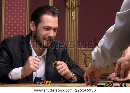 Handsome  confident man   in casino looking at  chips smiling  sitting at table with  chips and cards waist up