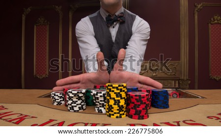 Handsome  dealer giving  red blue and shite chips in piles  on  table with  in casino looking at camera