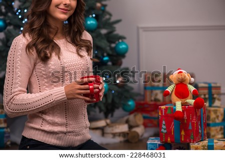 Beautiful attractive brown haired girl sitting near red Christmas socks for presents and fir tree and heap of  presents  on white pile carpet dressed in beige knitted jacket