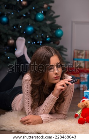 Beautiful attractive sad  disappointed brown haired girl laying on stomach with phone near  fir tree and heap of  presents  in different  gift wrapping on white pile carpet