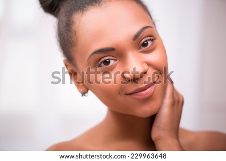 Portrait of beautiful  dark skinned girl in white towel with black curly hair in bun smiling touching her neck  looking at  camera isolated on white background