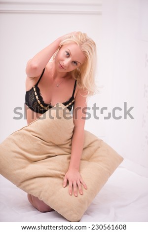 Sexy beautiful blond girl in black underclothes with blue eyes sitting on bed  with beige pillow looking at camera  isolated on white background
