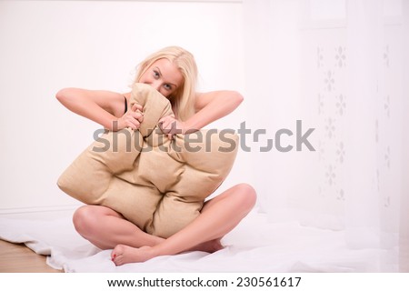 Sexy beautiful blond girl in black underclothes with blue eyes sitting looking out beige pillow and squeezing it  looking camera isolated on white background   with copy place