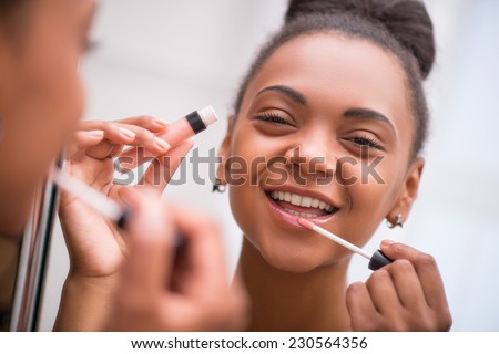 Beautiful  dark skinned girl making up her lips with rose lipstick or lip gloss isolated on white background smiling looking at mirror selective focus