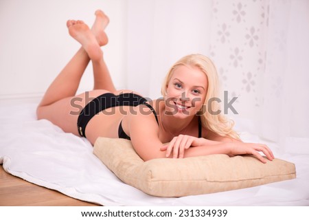 Sexy beautiful blond girl in black underclothes with blue eyes smiling  lying on stomach   on white spread on beige pillow looking camera  isolated on white background