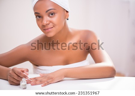 Portrait of beautiful  dark skinned girl in white towel on head and body painting her nails with transparent enamel looking at camera sitting at white table