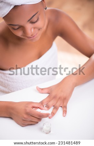 Portrait of beautiful  dark skinned girl in white towel on head and body painting her nails with transparent enamel looking at hands sitting at white table