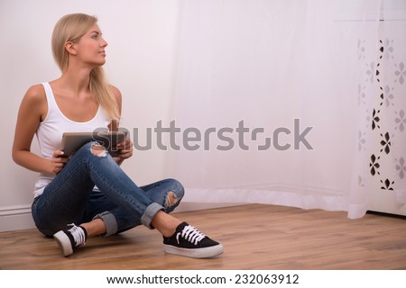 Beautiful happy  blond girl in white  T shirt  and jeans and trainers smiling looking aside  sitting on floor with  tablet computer with copy place full length