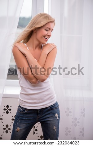 Beautiful happy  blond girl in white  T shirt  and jeans and trainers smiling looking aside touching her shoulders with hands waist up