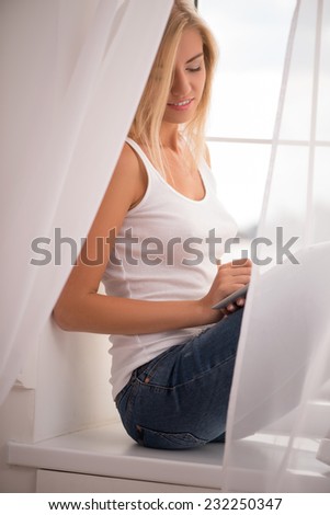Beautiful happy  blond girl in white  T shirt  and jeans smiling  sitting on windowsill looking at  tablet computer with copy place  waist up