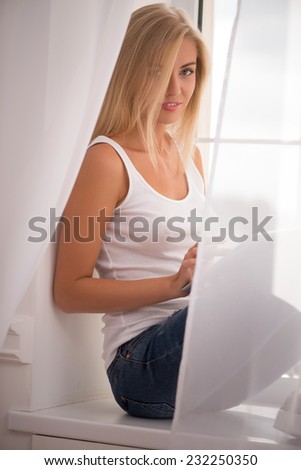 Beautiful happy  blond girl in white  T shirt  and jeans smiling  sitting on windowsill with  tablet computer looking at camera with copy place waist up