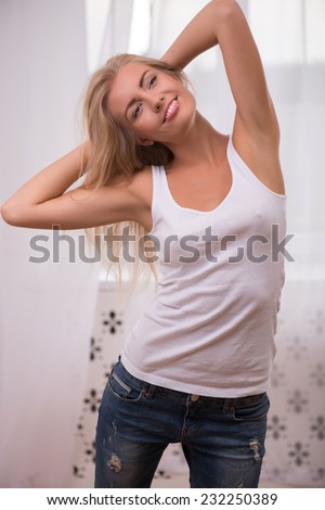 Beautiful happy  blond girl in white  T shirt  and jeans and trainers smiling looking aside holding her hands behind her head  waist up