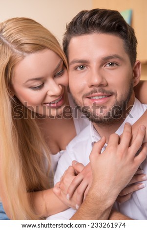 Portrait of happy couple in love of handsome man looking at camera and attractive woman  looking at him embracing  and him sitting on sofa