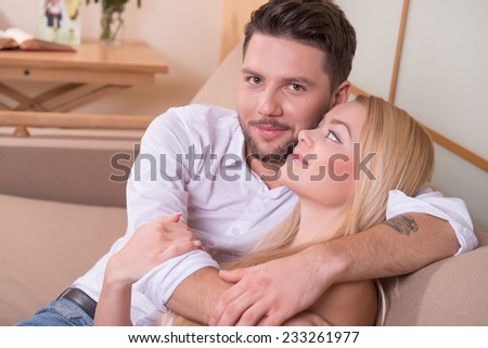 Portrait of happy couple in love of handsome man looking at camera and attractive woman looking at him sitting on sofa embracing smiling