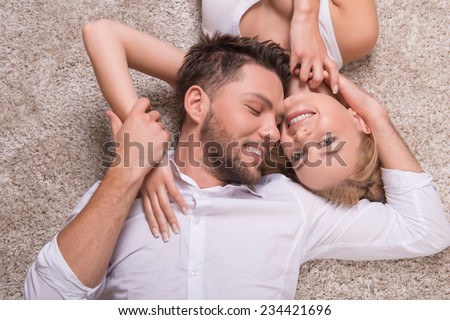 Portrait of happy couple in love of handsome man looking at attractive woman looking at camera smiling embracing  lying on carpet