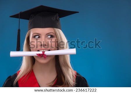 Portrait of beautiful happy blond girl in black cloak and master hat holding in mouth bundle of sheet of paper on blue background smiling looking aside