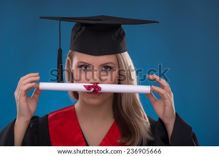 Portrait of beautiful happy blond girl in black cloak and master hat holding bundle of sheet of paper on blue background smiling looking at camera