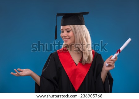 Portrait of beautiful happy blond girl in black cloak and master hat holding bundle of sheet of paper on blue background smiling looking aside
