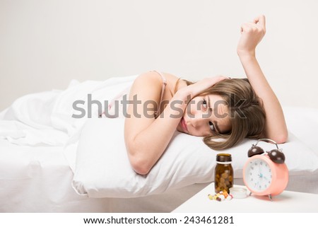Insomnia. Closeup of young beauty lying in bed with pills and clock on the table and looking up suffering from insomnia
