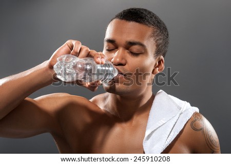 Refreshing after training. Young shirtless African man holding bottle with water and drinking while standing against grey background with copy space