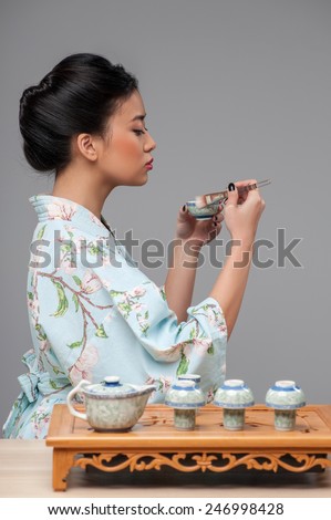 Japanese tea ceremony. Side view image of young beautiful Japanese woman in traditional kimono holding cup of tea and cleaning it while standing by tea table against grey background